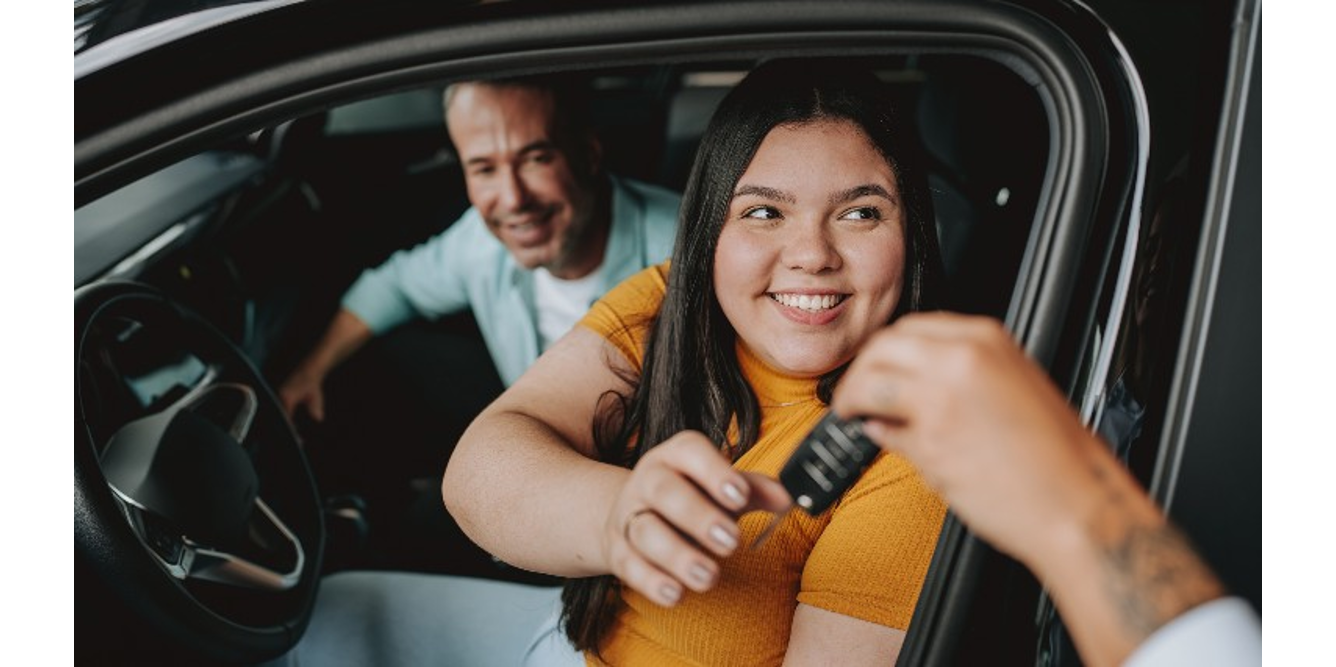 Smiling young woman in yellow top receiving car keys while sitting in driver's seat, with a happy man looking on through the passenger window.