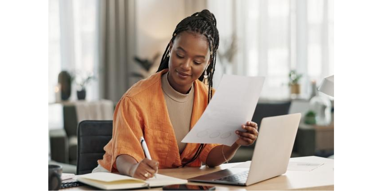 Young woman reviewing a document while taking notes at her desk with a laptop in a bright home office.