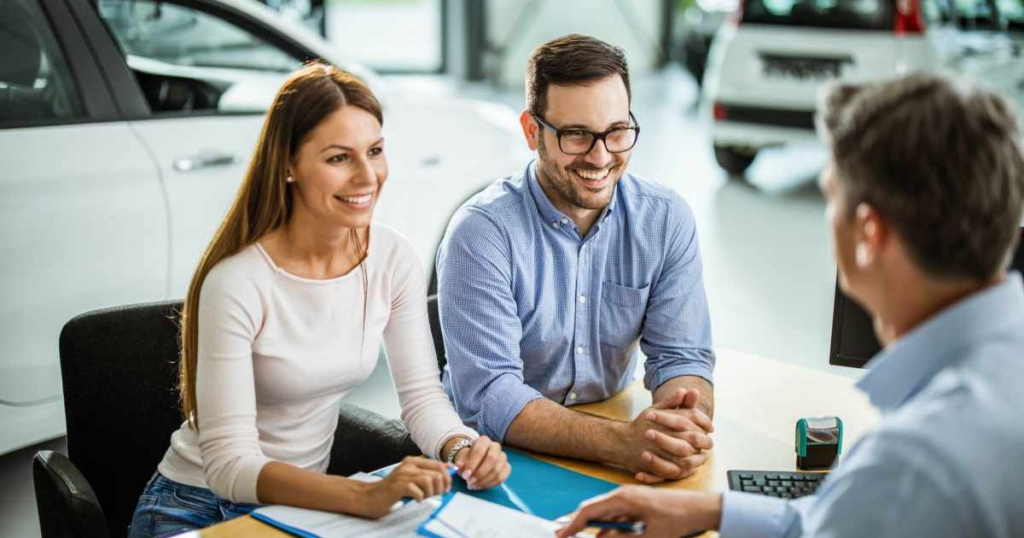 A Happy couple sitting at a desk with a car dealership salesperson, reviewing paperwork to purchase a vehicle in a showroom.