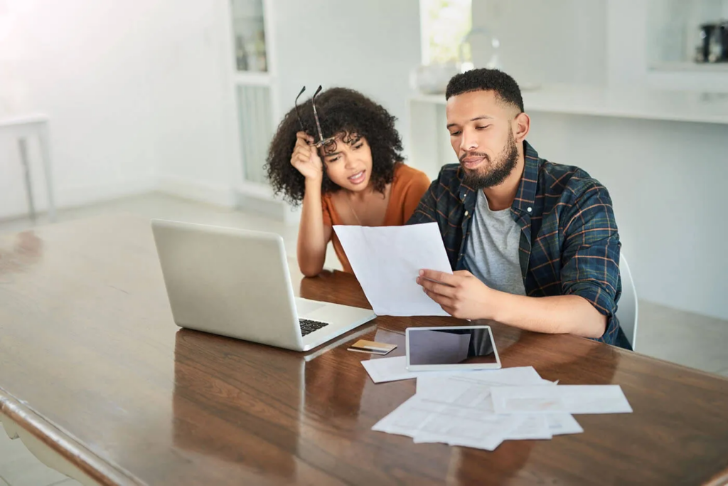 Couple reviewing bills and paperwork at their kitchen table with a laptop and tablet.